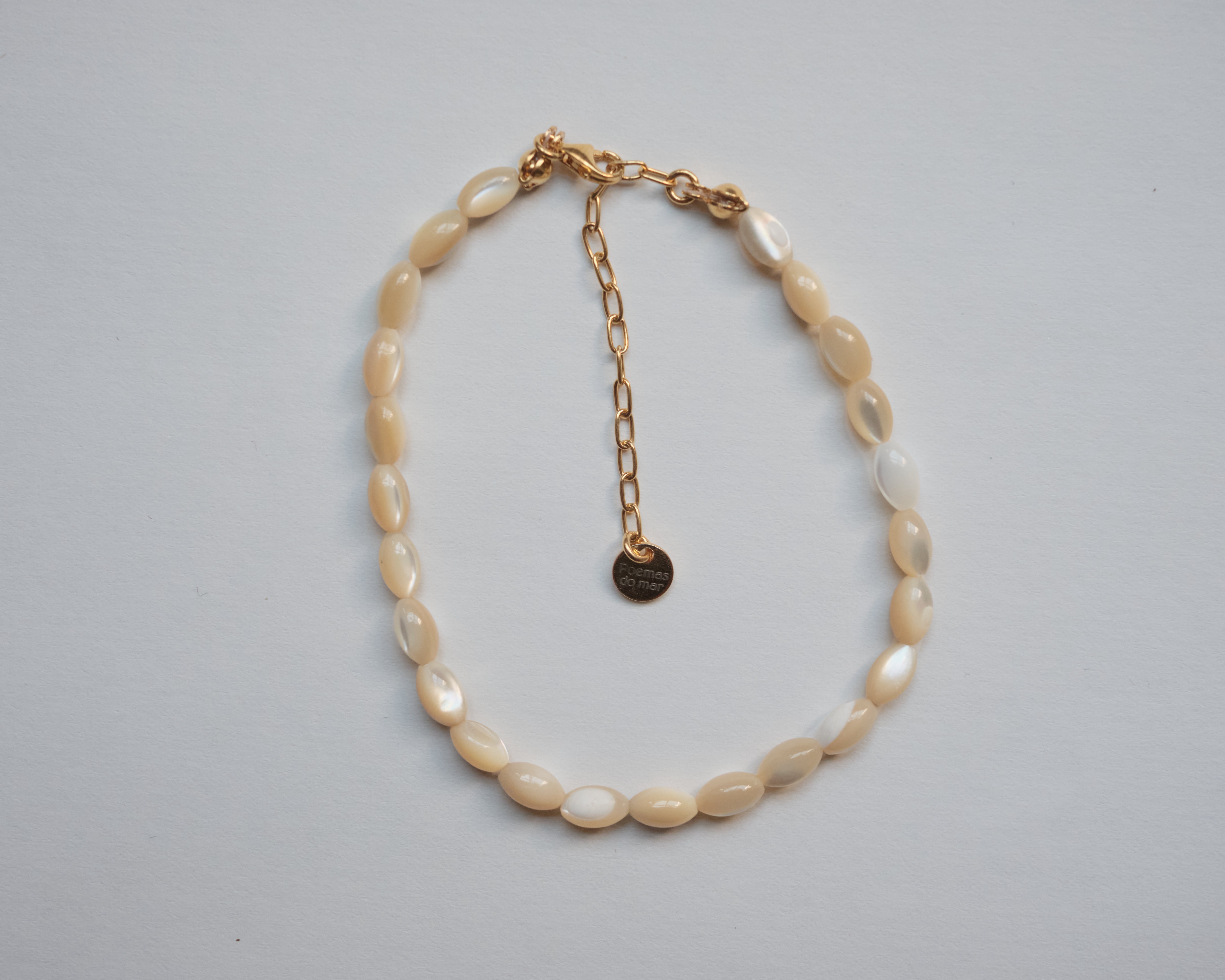 A beaded necklace made of irregular cream-colored shell-like beads with a gold-tone chain extender and small round tag, photographed on a plain light gray background.