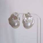 A pair of large, irregular baroque pearl stud earrings with a pearlescent white and soft gray luster, displayed on a clear acrylic stand against a neutral light gray background.