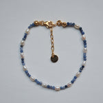 A delicate beaded bracelet with white baroque pearls, light blue faceted beads, and gold-toned metal hardware, including a lobster clasp, extender chain, and round tag, displayed on a light gray background.