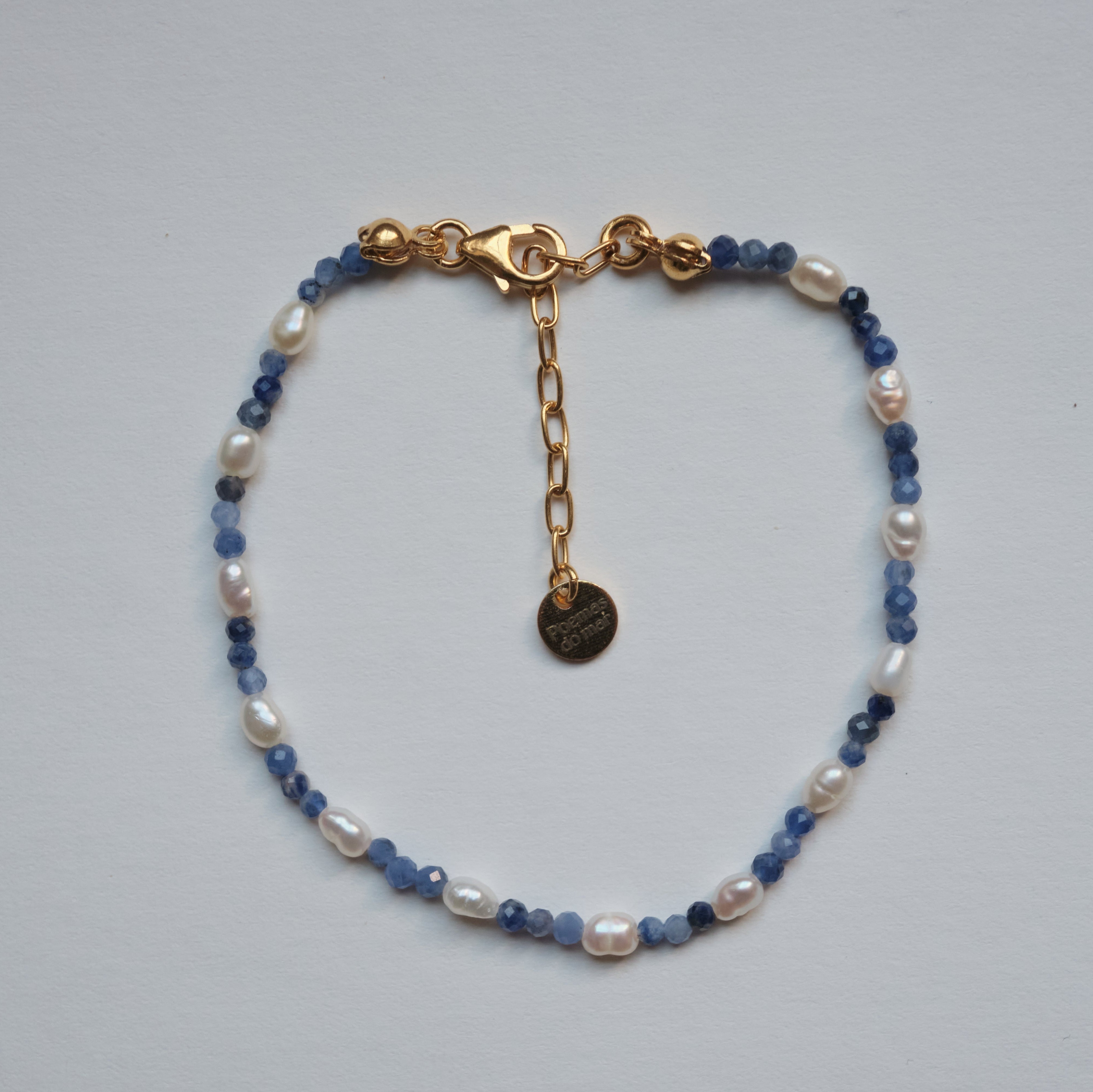 A delicate beaded bracelet with white baroque pearls, light blue faceted beads, and gold-toned metal hardware, including a lobster clasp, extender chain, and round tag, displayed on a light gray background.