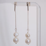 A pair of long dangling earrings with silver-toned chains, a small round pearl, and a larger irregular baroque pearl, hanging from a clear display stand against a light gray background.