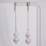 A pair of long dangling earrings with silver-toned chains, a small round pearl, and a larger irregular baroque pearl, hanging from a clear display stand against a light gray background.