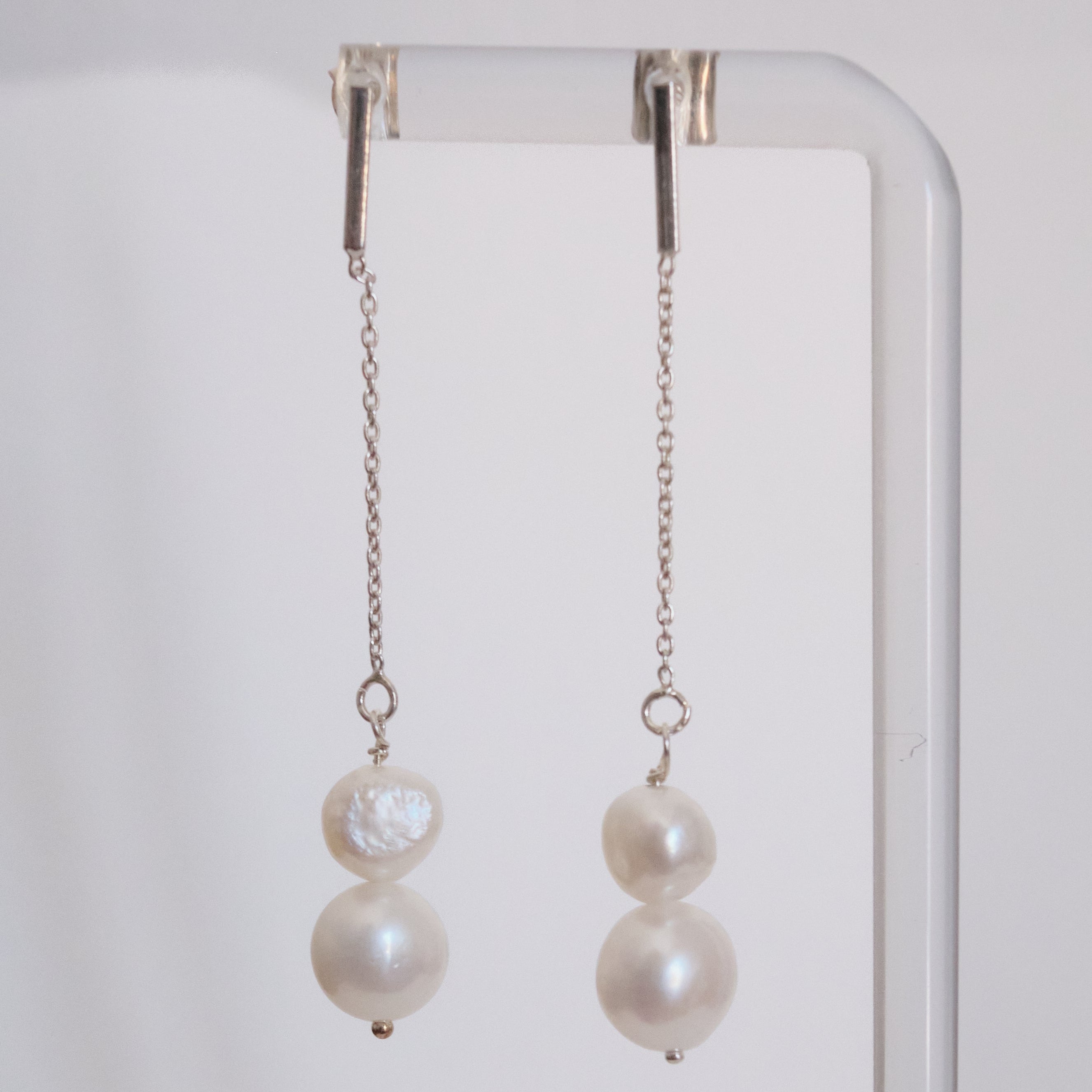 A pair of long dangling earrings with silver-toned chains, a small round pearl, and a larger irregular baroque pearl, hanging from a clear display stand against a light gray background.