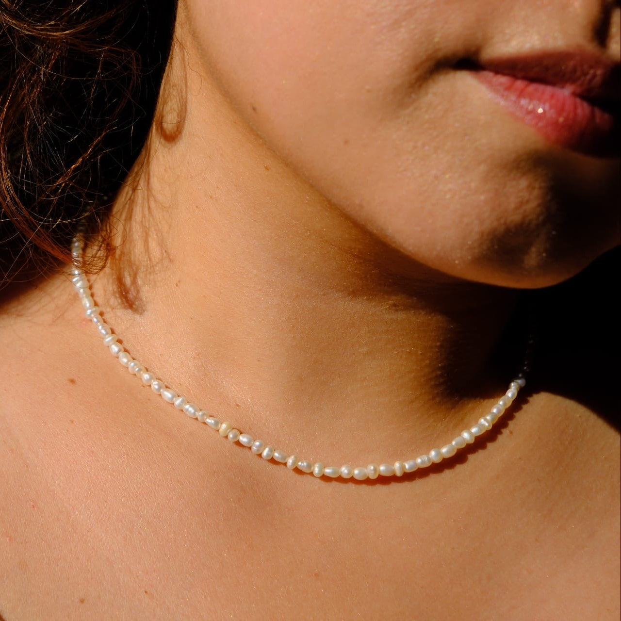 A close-up lifestyle shot of a woman wearing a delicate white pearl necklace, showing her neck, collarbone, and lower face in warm sunlight.