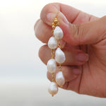 A hand holding a pair of gold drop earrings with irregular white baroque pearls, set against a soft, blurred outdoor background.