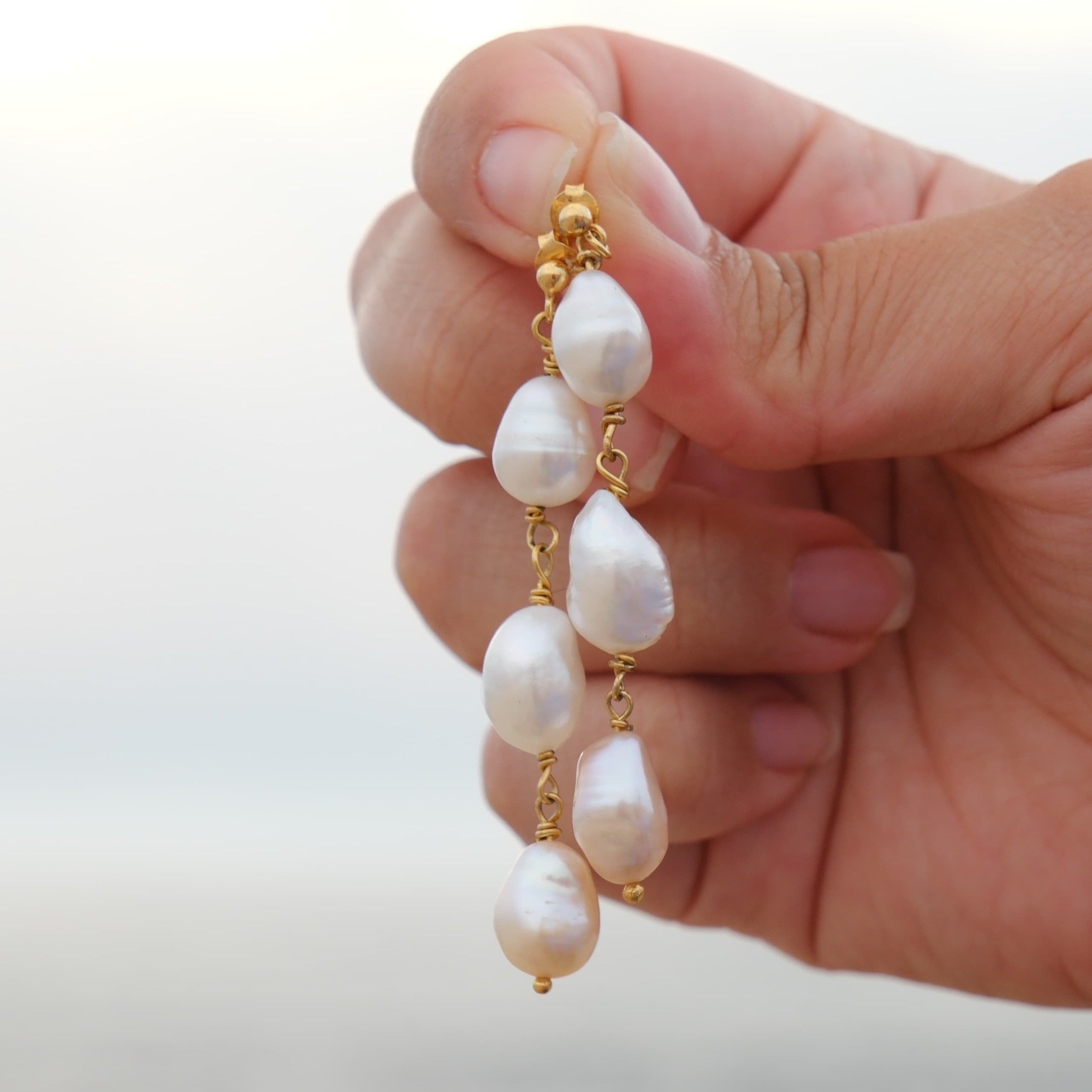 A hand holding a pair of gold drop earrings with irregular white baroque pearls, set against a soft, blurred outdoor background.