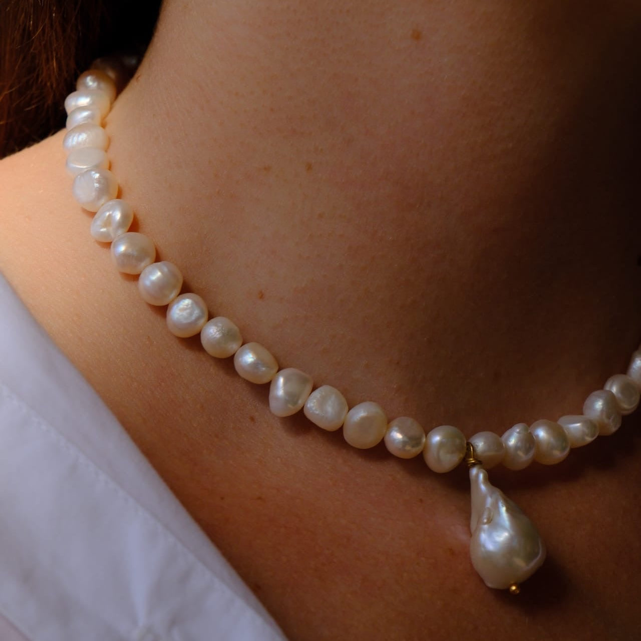 A close-up of a person's neck wearing a baroque pearl necklace with a teardrop-shaped pearl pendant and a small gold accent.