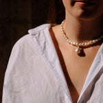 A close-up lifestyle shot of a woman wearing a white linen shirt, a pearl necklace with a teardrop pendant, and matching pearl drop earrings, set against a dark, shadowed background.