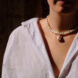 A close-up lifestyle shot of a woman wearing a white linen shirt, a pearl necklace with a teardrop pendant, and matching pearl drop earrings, set against a dark, shadowed background.