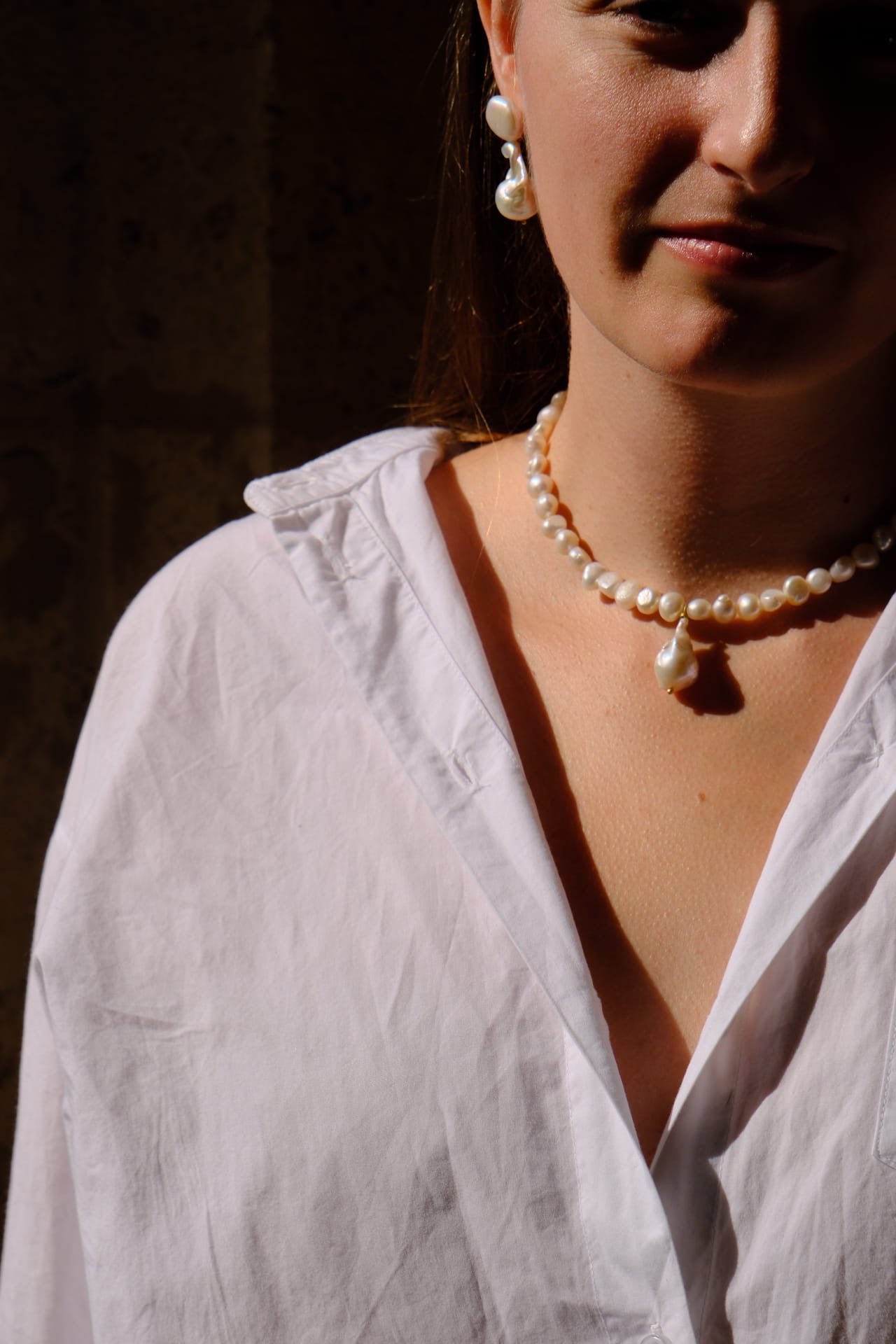 A close-up lifestyle shot of a woman wearing a white linen shirt, a pearl necklace with a teardrop pendant, and matching pearl drop earrings, set against a dark, shadowed background.