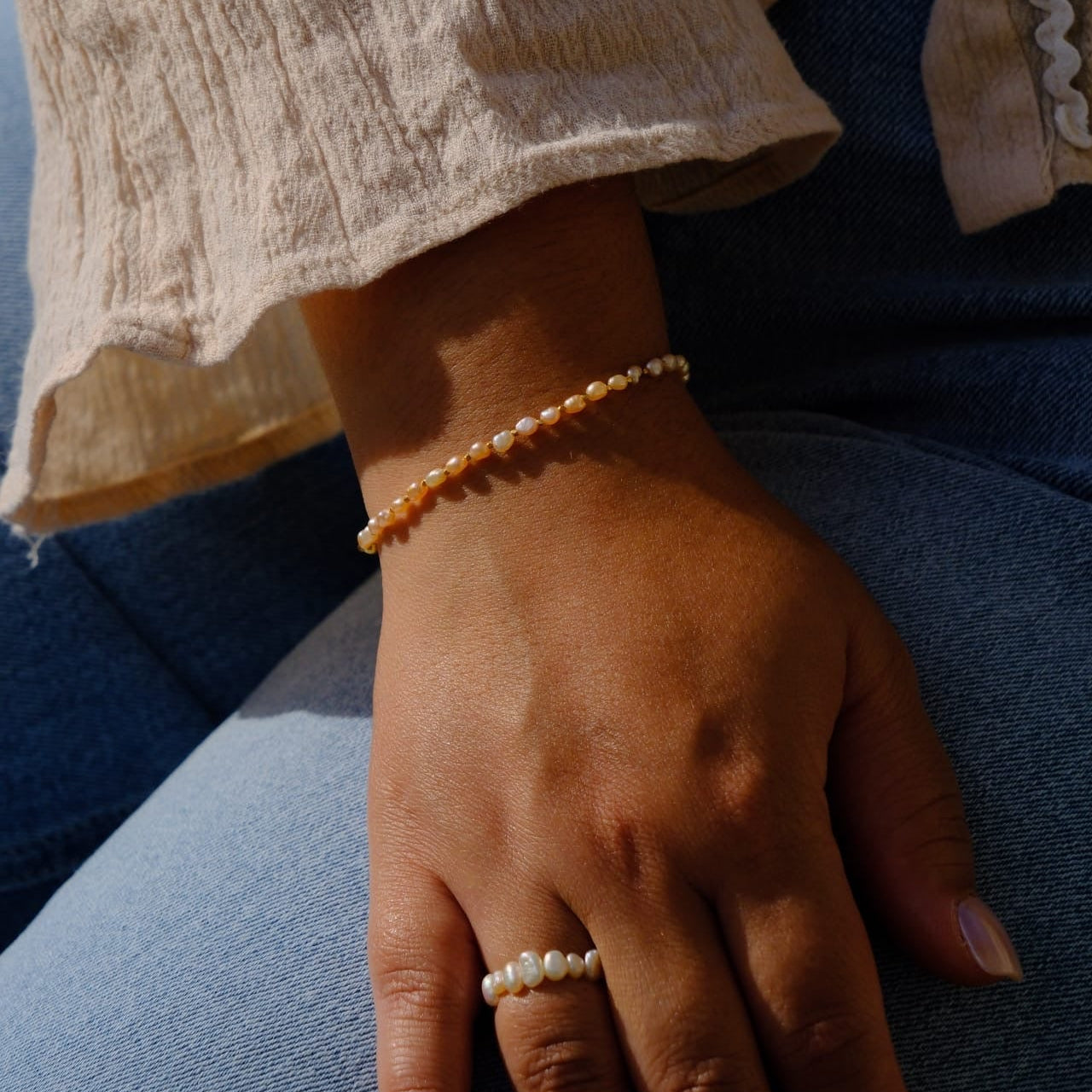 A close-up lifestyle shot of a person's hand resting on their lap, wearing a delicate gold beaded bracelet on the wrist and a matching gold beaded ring on the finger. The person is wearing a beige textured blouse with ruffled cuffs and blue jeans.
